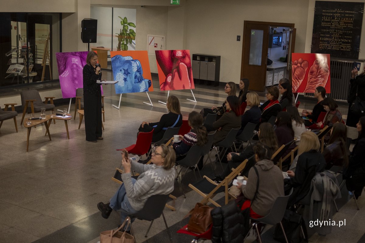 Kobieta stoi i przemawia do zebranej publiczności, w tle na sztalugach znajdują się trzy wielkoformatowe ilustracje gdyńskiej artyski.