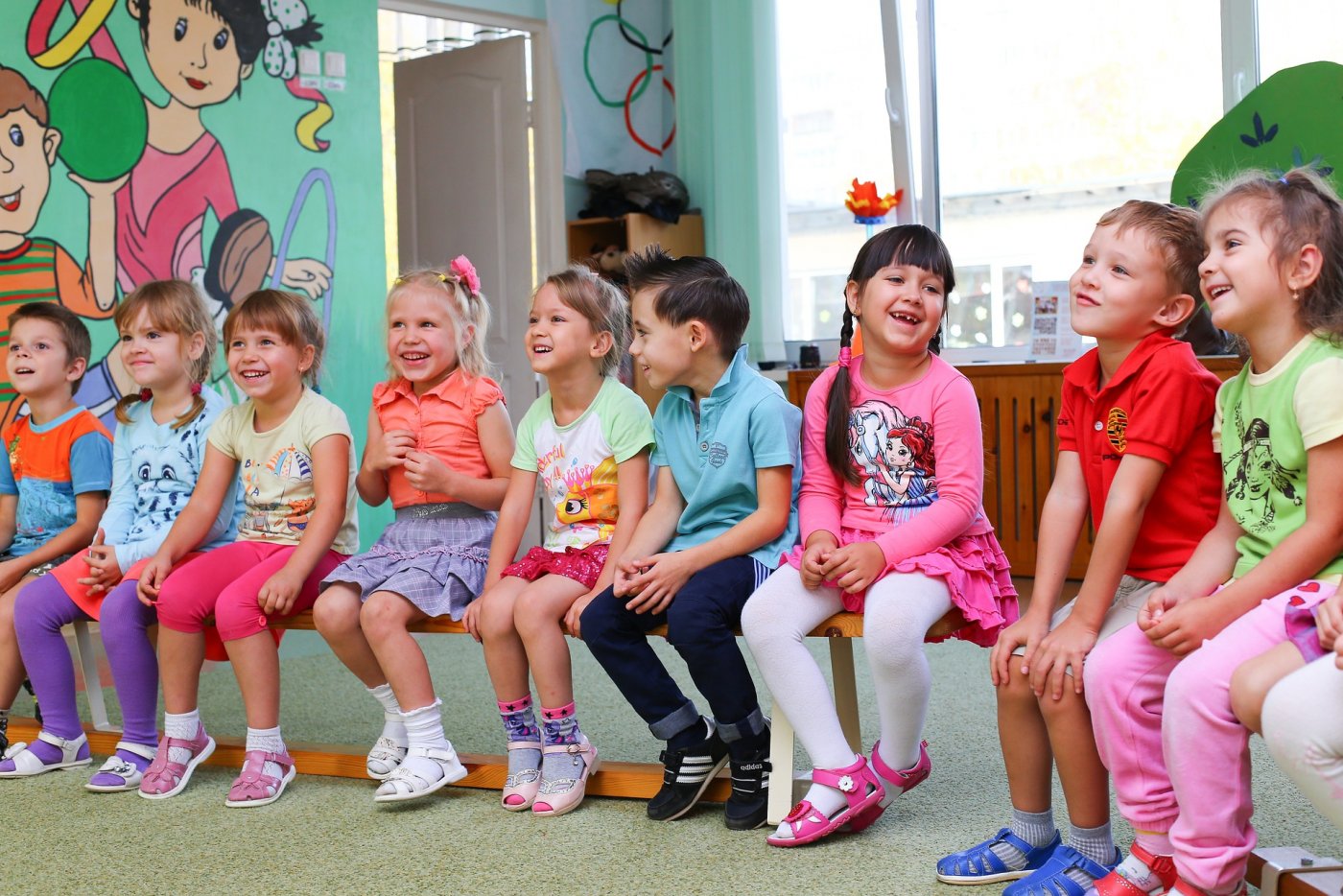 Na zdjęciu znajdują się dzieci w wieku przedszkolnym, siedzące na krzesełkach i uśmiechające się, ubrane w różnokolorowe ubrania. W tle znajduje się kolorowa sala przedszkolna.