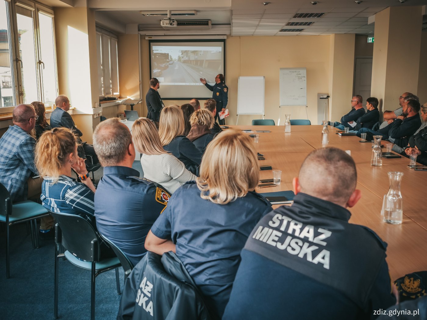 spotkanie w sali, widoczne osoby siedzące przy stole, na pierwszym planie jest osoba siedząca tyłem na napisem na bluzie:Straż Miejska. W tle trwa prezentacja.