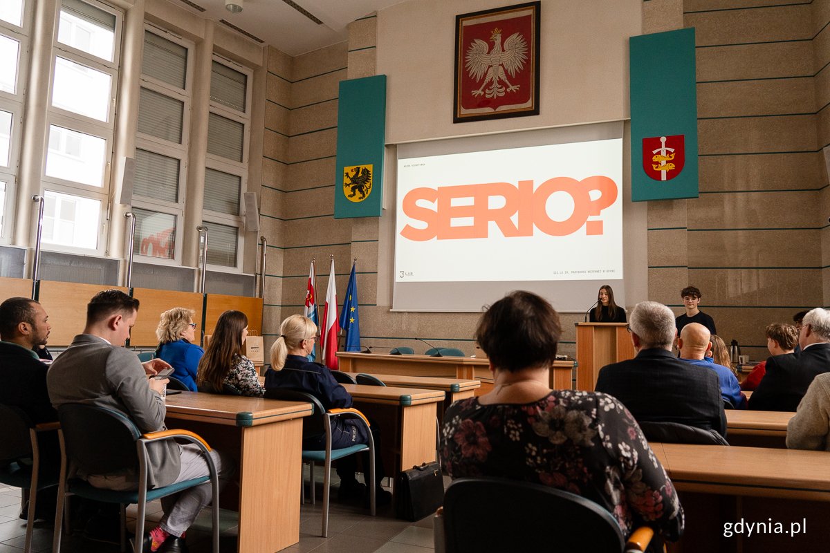 Uczniowie z III LO w Gdyni występują przed publicznością w sali Rady Miasta Gdyni. Na ekranie wyświetlona jest plansza z dużym napisem 