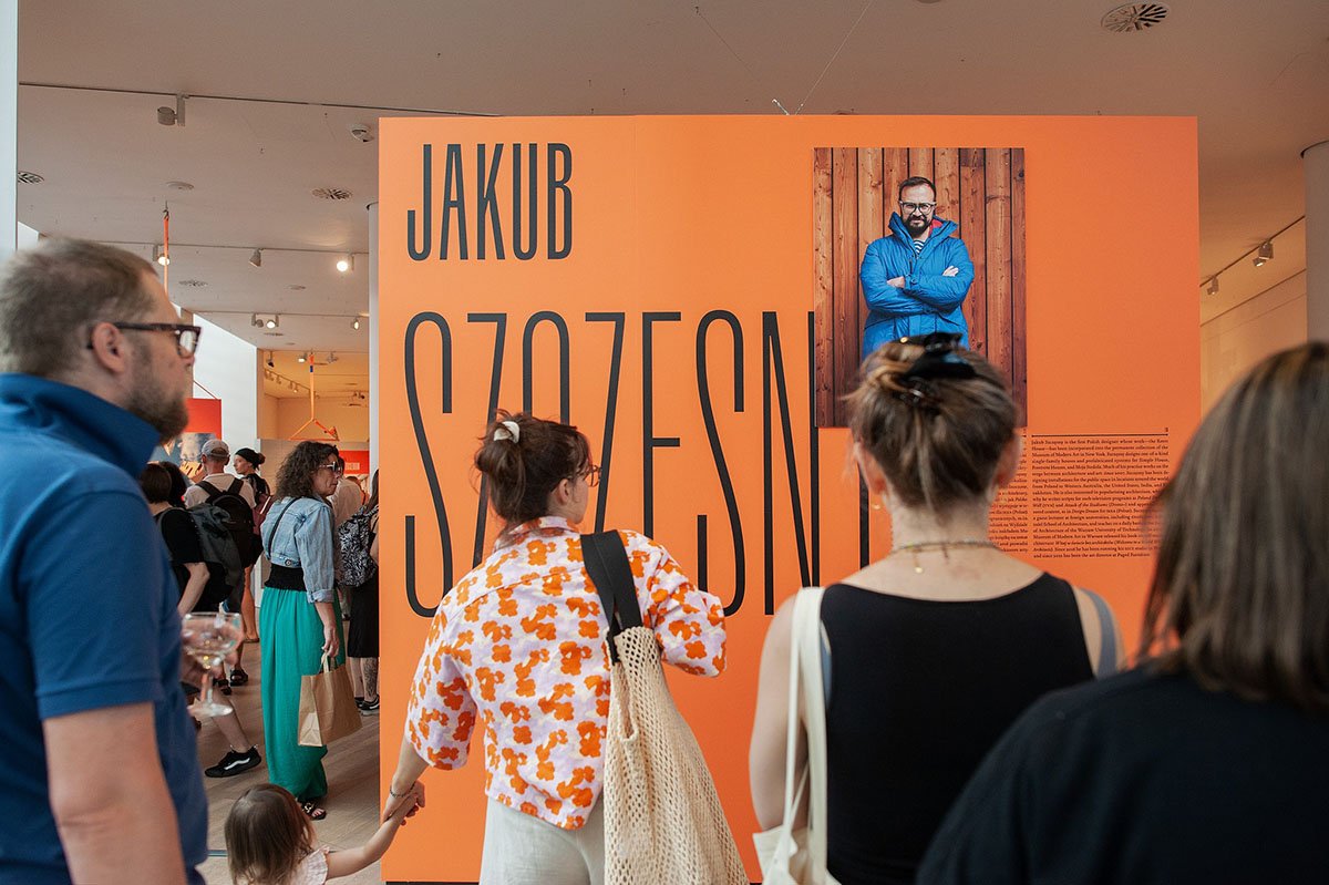 ludzie przed banerem wystawy Jakuba szczęsnego. Na banerze zdjęcie uśmiechniętego mężczyzny w niebieskiej kurtce