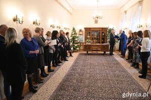 W sali Urzędu Stanu Cywilnego stoją po obu stronach nauczycielki i zaproszeni goście w centrum kadru masywne drewniane meble, na środku wzorzysty dywan, na ścianach wiszą kandelabry