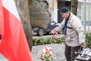 Mieszkaniec kładzie na pomniku biało-czerwoną wstęgę