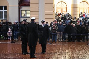 Na pierwszym planie znajdują się trzej mężczyźni w mundurach, na drugim zgromadzona publiczność cywilna przy budynku AMW.