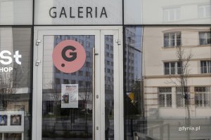 Przeszklona witryna Gdańskiego Centrum Filmowego wraz z szklanymi drzwiami z plakatem warsztatów. Nad drzwiami duży czarny napis galeria.