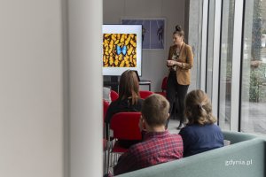 Na pierwszym planie siedzący uczestnicy warsztatów, na drugim planie kobieta przemawiająca do grupy, obok ekran z wyświetlonymi materiałami szkoleniowymi.