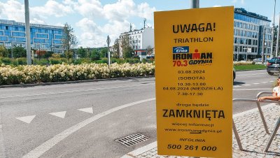 na pierwszym planie tablica informująca o zmianach w organizacji ruchu podczas imprezy sportowej, w tle miasto, ruch uliczny, ulica, zieleń i budynki