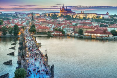 Popołudniowo-wieczorna panorama czeskiej Pragi. Na pierwszym planie po lewej stronie Most Karola pełen turystów. W dole mostu rzeka Wełtawa. Za mostem widoczne kamienice oraz zamek na wzgórzu.