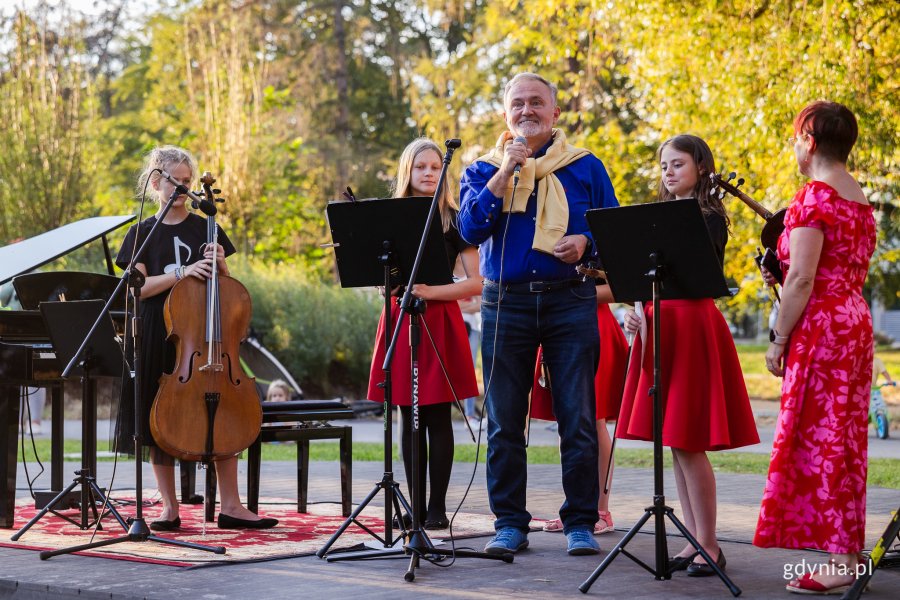 Mężczyzna mówi do mikrofonu w parku, obok stoją uczennice z instrumentami muzycznymi.