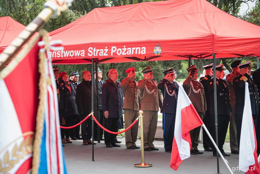 Uczestnicy obchodów Dnia Strażaka stoją pod czerwonym namiotem. 