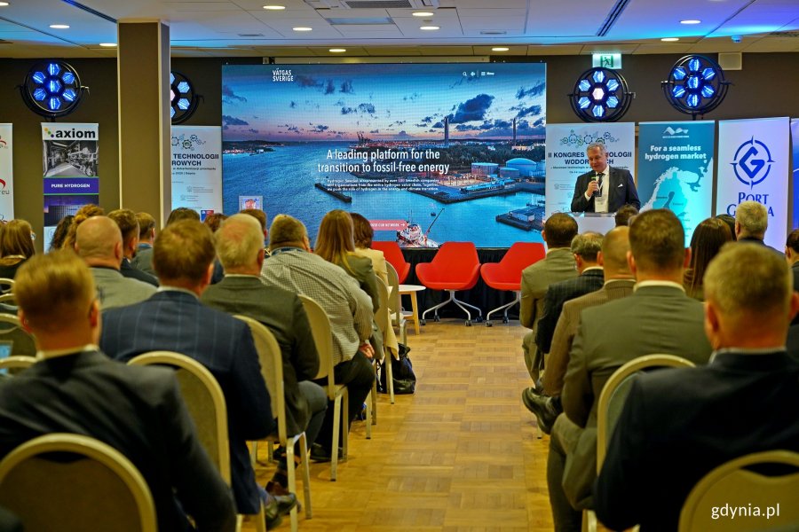 Na pierwszym planie: uczestnicy kongresu siedzący na krzesłach. Na drugim planie, przy mównicy: Paweł Seremak, head of projects w Vätgas Sverige & Baltic Sea Region Hydrogen Council, podczas wystąpienia na kongresie