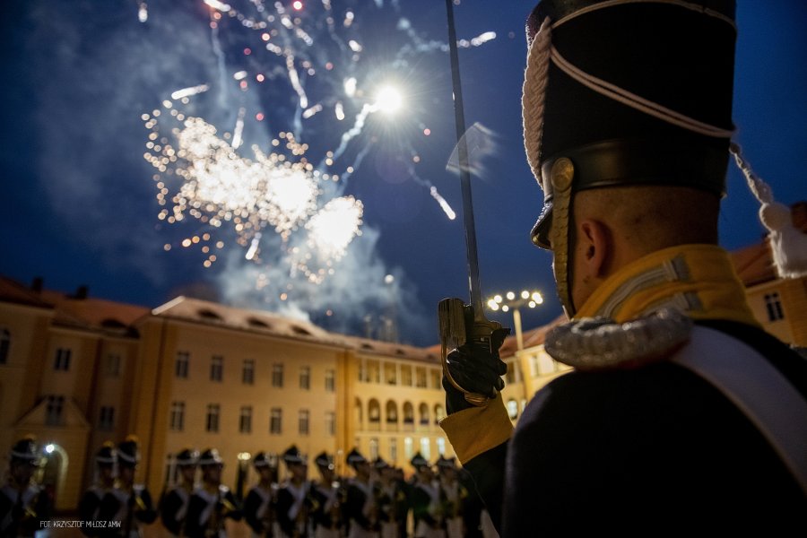 Podchorążowie w historycznych mundurach stoją na placu apelowym, nad nim wybuchaja fajerwerki.