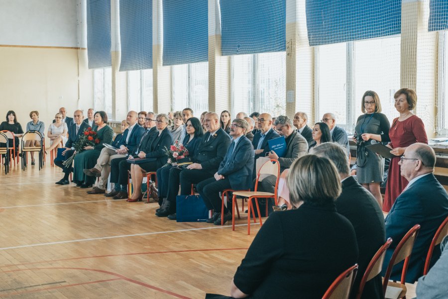 Publiczność wydarzenia siedząca na sali gimnastycznej, za nią okna z niebieskimi roletami