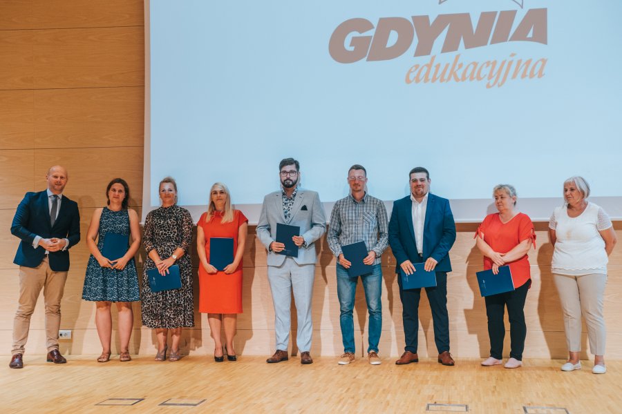 Na scenie w rzędzie grupa ludzi, w tle ekran rzutnika z logiem Gdyni edukacyjnej na białym tle 