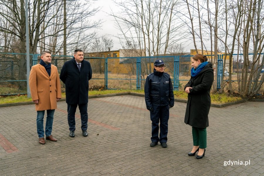 Dwóch mężczyzn, kobieta i policjantka stoją na parkingu, w tle ogrodzenie i tereny przemysłowe