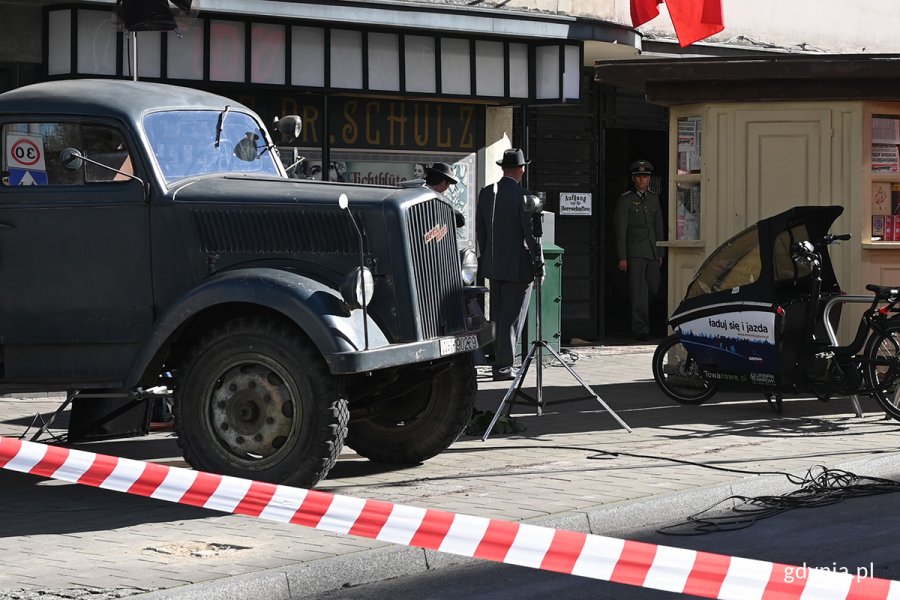 Trzy osoby w strojach z lat 40. rozmawiające w wejściu do budynku pomiędzy ciężarówką a kioskiem