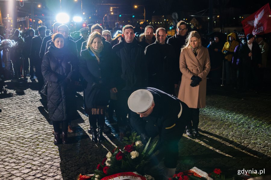 Żołnierz Marynarki Wojennej składa wieniec pod pomnikiem, za nim stoi grupa osób reprezentująca władze miasta.