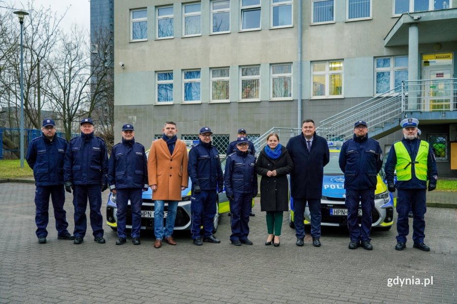 Grupa osób, w tym funkcjonariusze policji pozują do zdjęcia na tle radiowozów. W tle budynek komisariatu policji