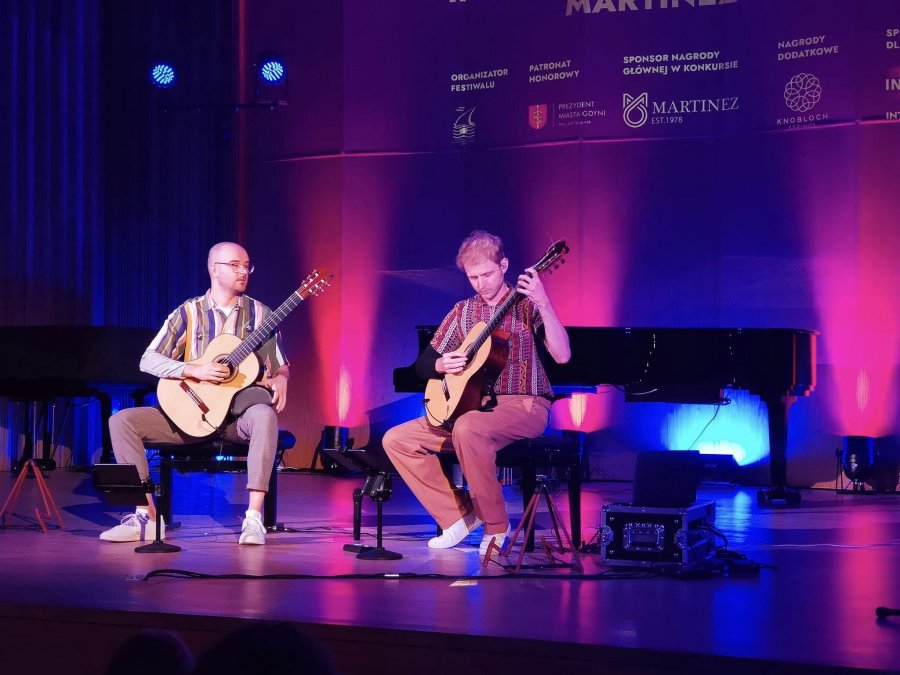 Dwóch mężczyzn na scenie, siedząc na krzesłach gra na gitarze, kolorowe oświetlenie