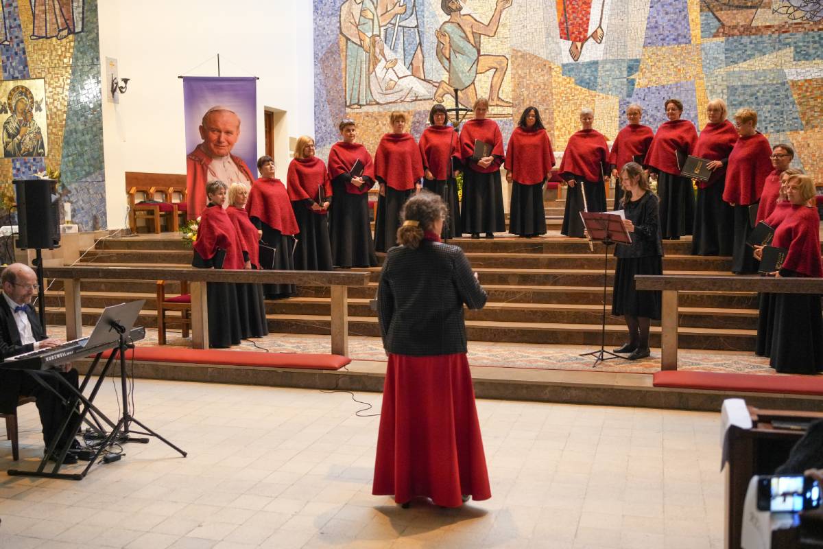 Kobiecy chór w czarno-czerwonych strojach stojący na schodach w kościele i mający za plecami witraż przedstawiający sceny biblijne. Obok widoczny muzyk z instrumentem klawiszowym