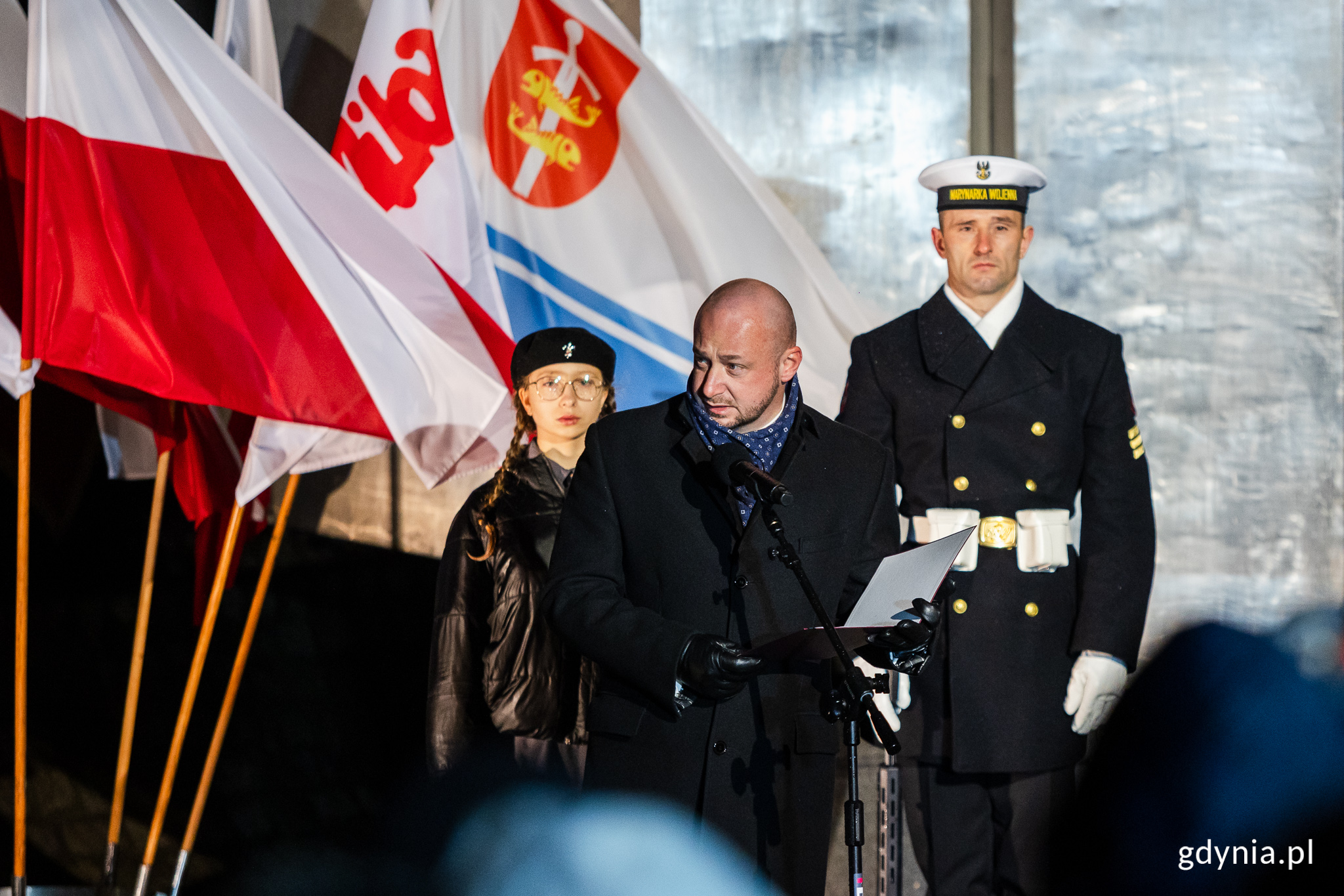 Mężczyzna przemawia do publiczności, w tle żołnierz Marynarki Wojennej i harcerka oraz pomnik Ofiar Grudnia '70