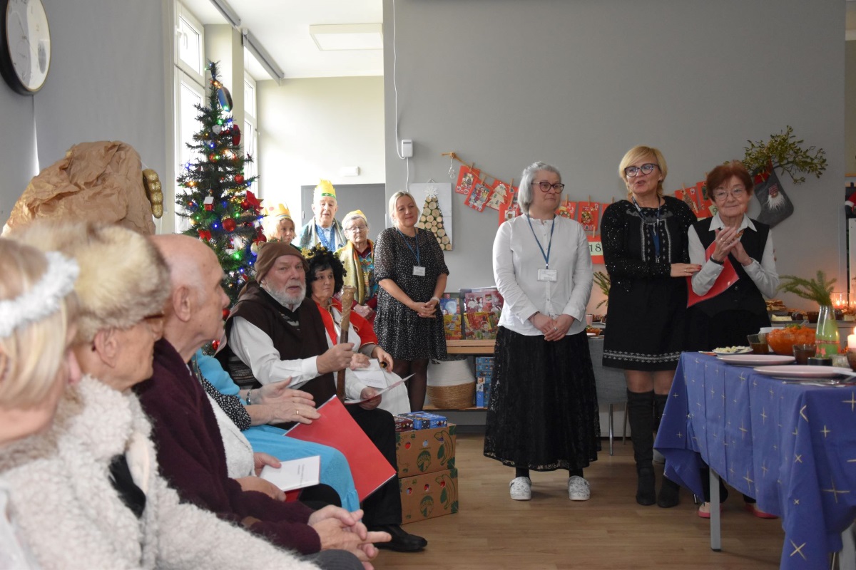 Zdjęcie przedstawia grupę osób w wieku senioralnym zaraz po wykonaniu przez nich autorskiej wersji jasełek. W tle widać kierownik domu obejmującą jedną seniorkę i dziękującą wszystkim za cudowny spektakl. 