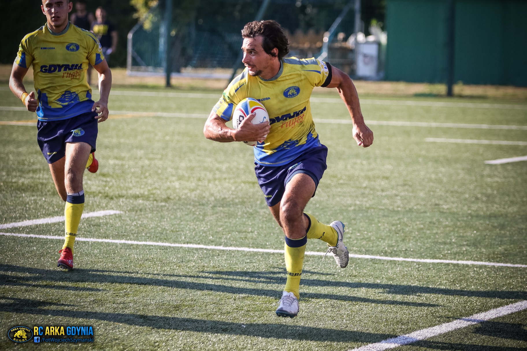 Na zdjęciu widzimy dwóch rugbystów Arki Gdynia. Obaj ubrani są w żółto-niebieskie stroje. Jeden z nich w prawą ręką trzyma jajowatą piłkę. Biegnie do przodu. Jego kolega z drużyny podążą za nim. Obaj znajdują się na pokrytym zielona murawą boisku // fot. Wojciech Szymański (Rugby Club Arka Gdynia)