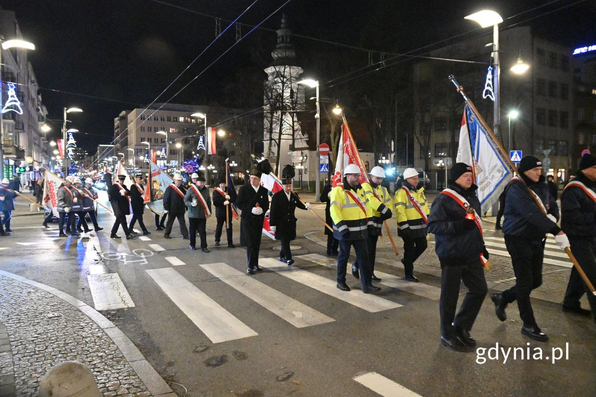 osoby maszerujące ulicą i niosące sztandary oraz znicze