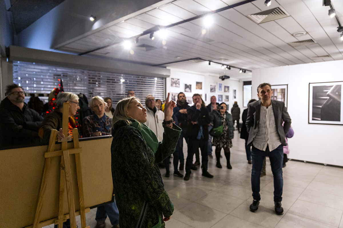 Kobieta w galerii sztuki opowiada o wystawianych pracach, w tle uczestnicy wernisażu fotografii.