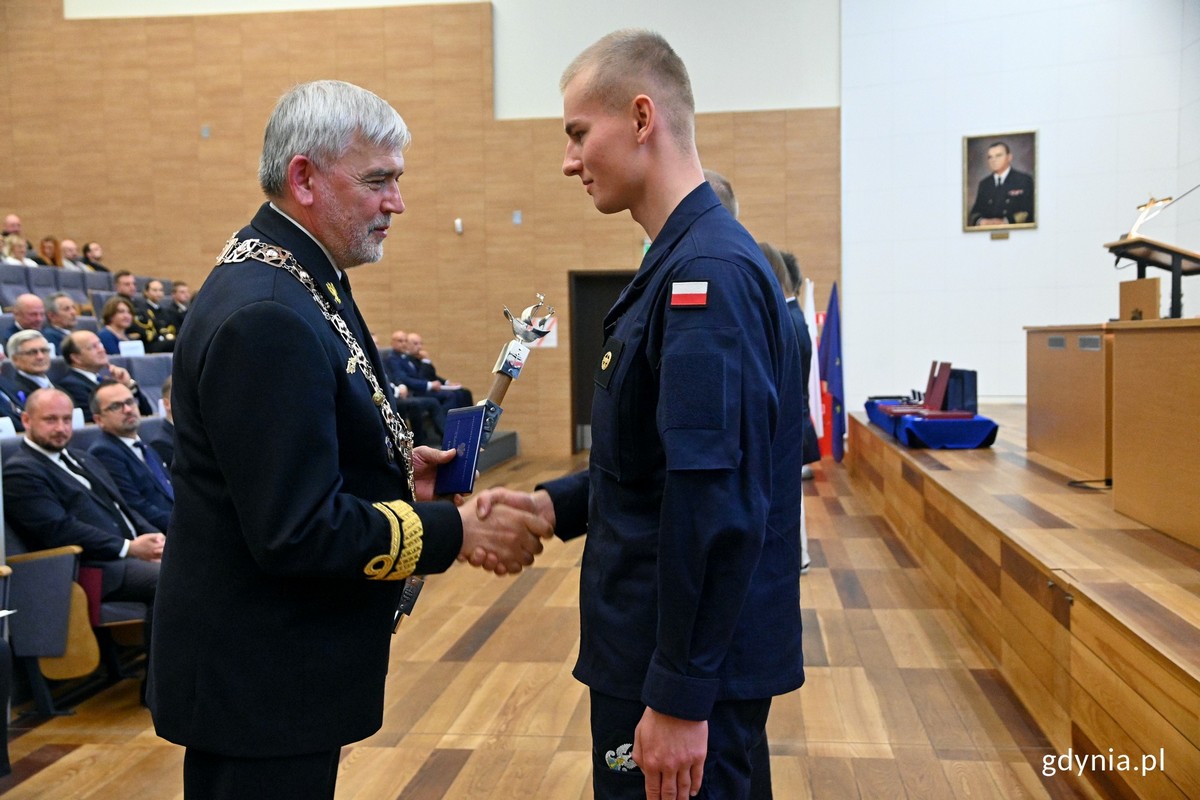 Sala w AMW, rektor uczelni ściska dłoń studenta w mundurze, w tle widownia pełna ludzi